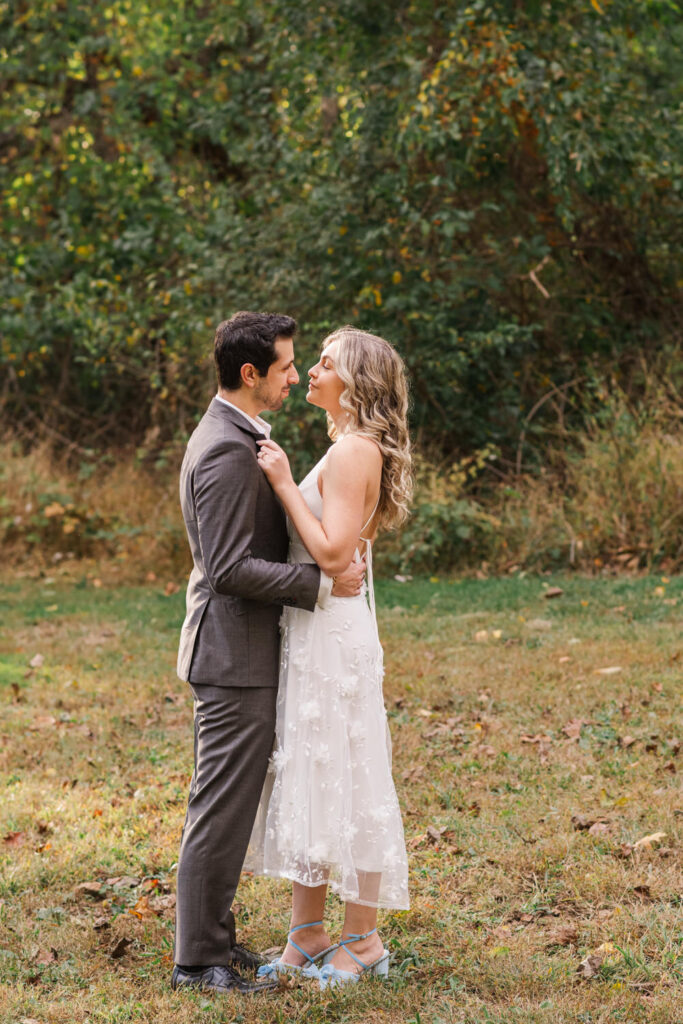 A couple embraces in a grassy area with trees in the background, the woman in a white dress and blue shoes, and the man in a dark suit, both gazing into each other's eyes.