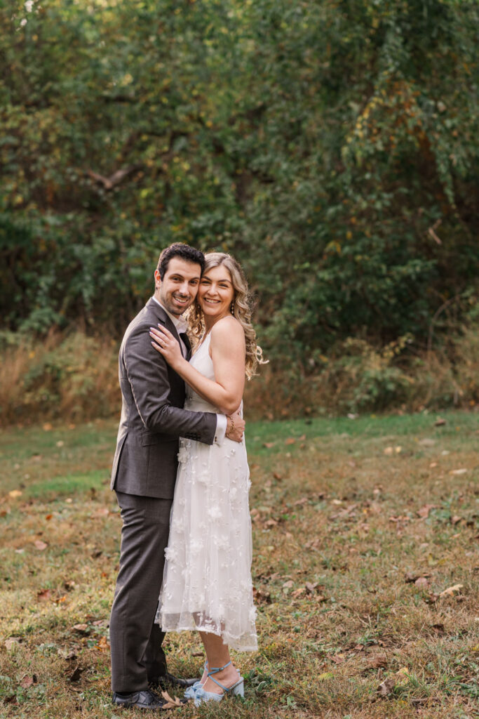 A couple embraces in a grassy area with trees in the background, the woman in a white dress with floral details and the man in a dark suit, both smiling at the camera.