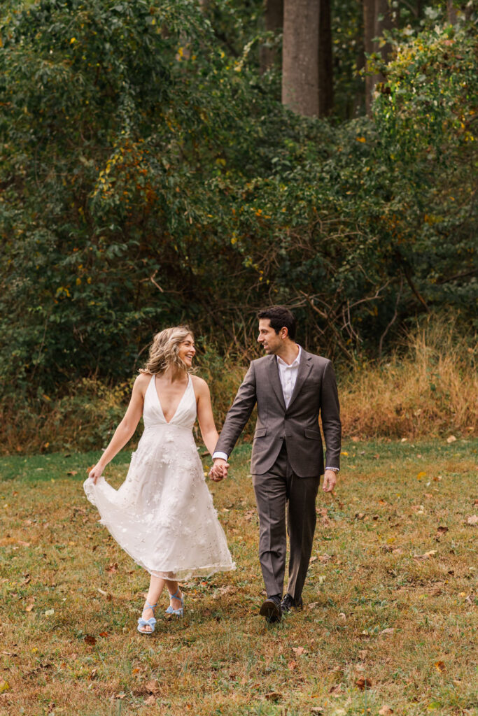 A couple holds hands while walking through a grassy field, the woman’s white dress flowing as she smiles at the man in a dark suit.