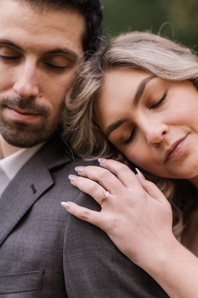 Close-up of the couple with their eyes closed, the woman resting her head on the man's shoulder, showcasing her engagement ring on his arm.