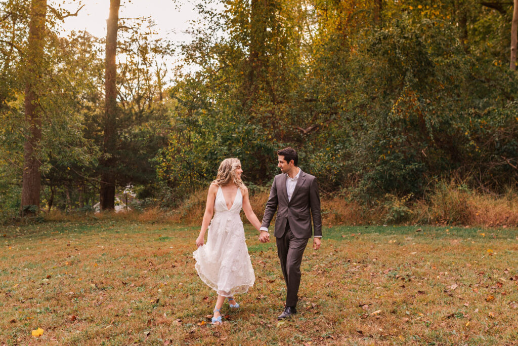 A couple walks hand-in-hand through a grassy field with trees in the background, the woman in a white dress and the man in a dark suit, both looking at each other.