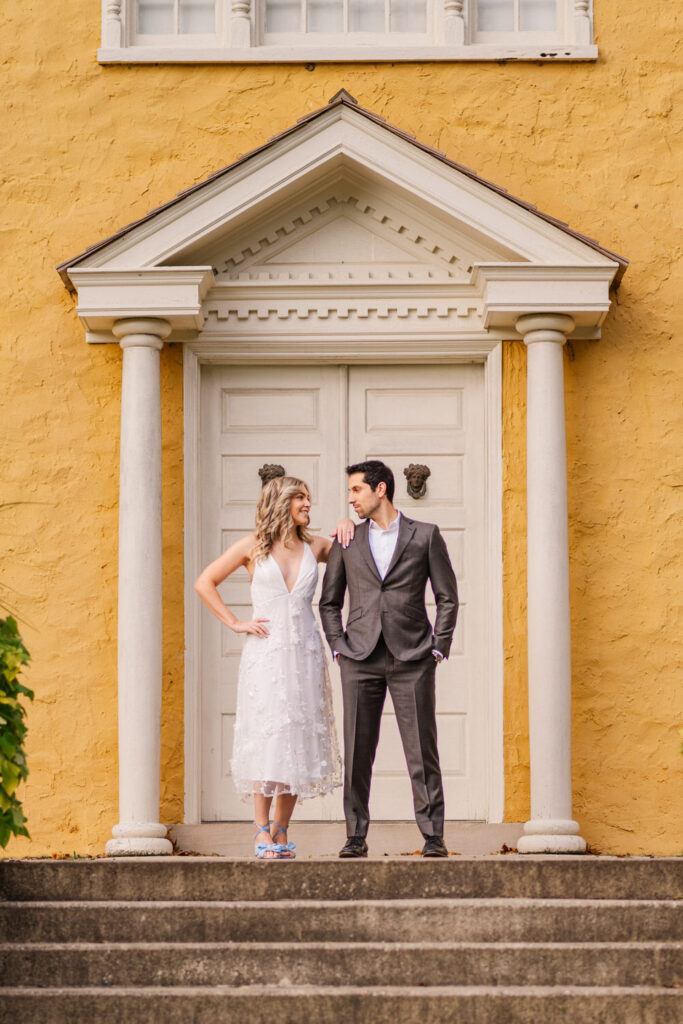 A couple stands on the steps of a historic building with a yellow exterior, the woman in a white dress with blue shoes and the man in a dark suit, both looking at each other with smiles.