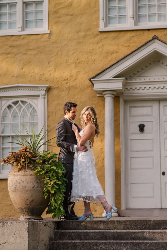 A couple poses on the steps of a historic building with a yellow exterior, dressed in semi-formal attire, the woman in a white dress and blue shoes, and the man in a dark suit.