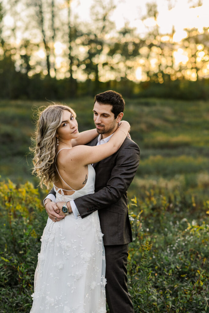 The couple embraces in a grassy field with the sun setting behind them, the woman in a white dress with an open back, and the man in a dark suit, both gazing into the distance.