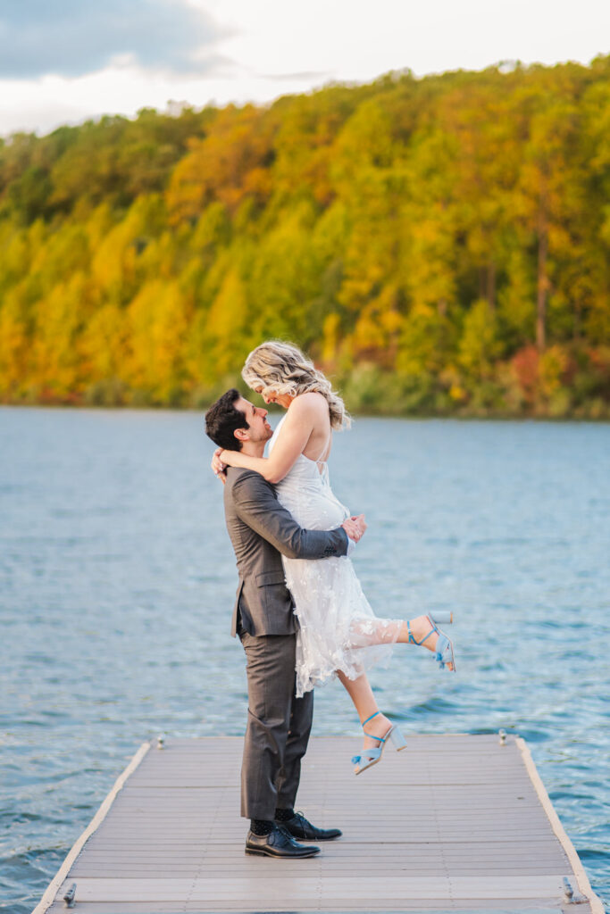 A man lifts the woman in a white dress as they stand on a dock by a lake, surrounded by autumn-colored trees, both smiling and looking at each other lovingly.