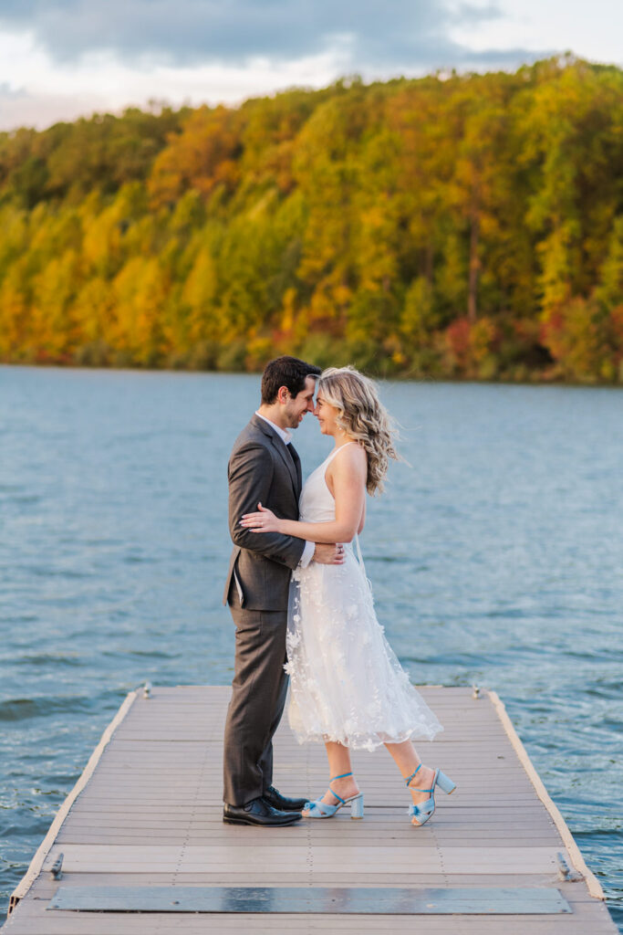 The couple stands closely on a dock by a lake, the woman in a white dress and blue shoes, and the man in a dark suit, both smiling and looking at each other.