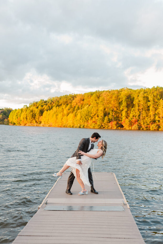 A man dips a woman in a white dress as they stand on a dock by a lake, both smiling at each other, surrounded by autumn foliage.