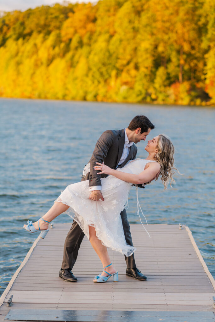 A man dips the woman in a white dress on a dock by a lake, surrounded by autumn-colored trees.