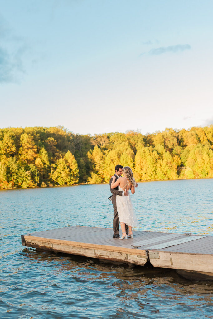 The couple dances on a dock by a lake, surrounded by autumn foliage, the woman in a white dress and blue shoes, and the man in a dark suit.