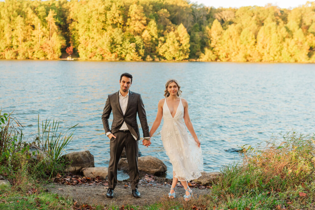 A couple holds hands by a lake, surrounded by autumn-colored trees, the woman in a white dress and the man in a dark suit, both looking at the camera.