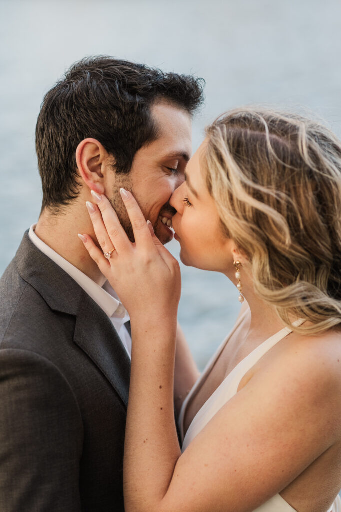 Close-up of the couple sharing an intimate kiss, the woman touching the man’s face, showcasing her engagement ring, set against a blurred outdoor backdrop.