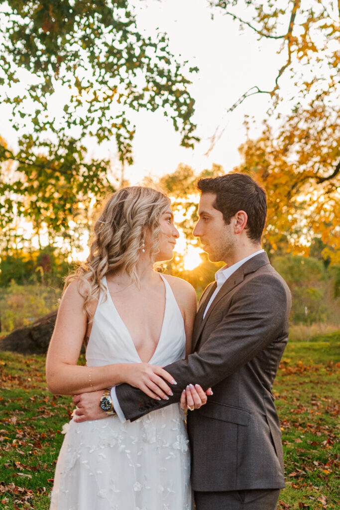 A couple stands closely together in a park with the sun setting behind them, the woman in a white dress and the man in a dark suit, both gazing into each other’s eyes.